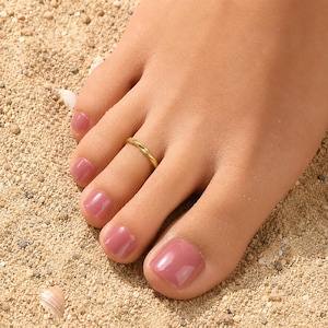 May include: A gold-toned metal toe ring featuring a simple, smooth, twisted design. The ring is worn on the second toe, which has a glossy, mauve-colored nail polish. The band is thin and circular, showcasing a polished, reflective metallic finish.