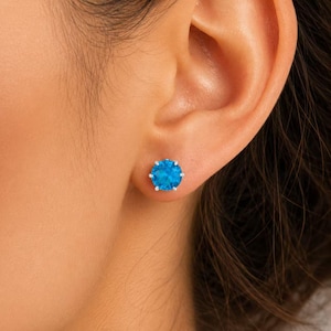 Könnte beinhalten: Ein runder Ohrstecker mit einem leuchtend blauen Edelstein im Brillantschliff, gefasst in eine silberfarbene Metallfassung mit Krappen. Der Ohrstecker sitzt mittig auf dem Ohrläppchen und zeigt eine kräftige, blaue Farbe mit Facetten.