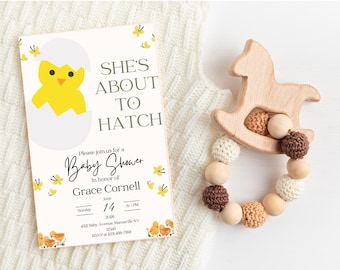 May include: A baby shower invitation with the text "She's About to Hatch" and a graphic of a yellow chick hatching from an egg. A wooden rocking horse-shaped baby rattle with knitted and wooden beads is next to the invitation.