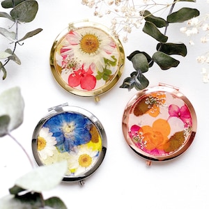 May include: Three circular compact mirrors, each containing pressed flowers. One has a gold frame with red and white blooms, another a silver frame with blue and white, and the last a rose gold frame with orange and pink flowers.