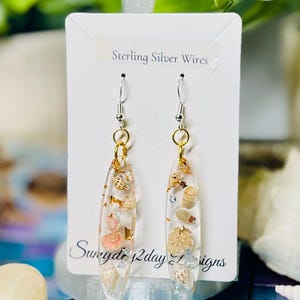 May include: Clear resin earrings with embedded seashells and gold flakes. The earrings have sterling silver wires and gold-colored hoops. The text on the card reads "Sterling Silver Wires" and "Swayde 2day Designs".