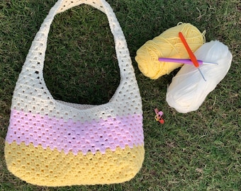 May include: A handmade crochet bag in cream, light pink, and yellow, with a matching strap. Two skeins of yarn, one yellow and one white, and two crochet hooks are next to the bag. A small pink flower is also visible.