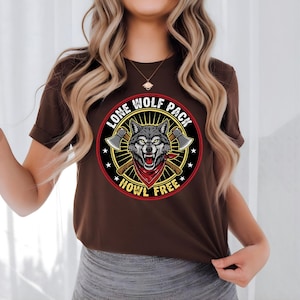 Puede incluir: Camiseta marrón con un diseño gráfico circular. El diseño presenta la cabeza de un lobo con un pañuelo rojo, hachas cruzadas y el texto "LONE WOLF PACK HOWL FREE". El diseño utiliza gris, rojo, negro y amarillo.