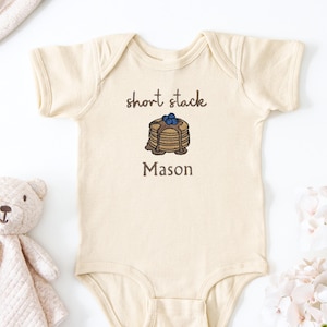 Puede incluir: Body de bebé color crema con las palabras "short stack" y "Mason" impresas. Una ilustración de panqueques con arándanos se encuentra entre el texto. Un osito de peluche y flores están en el fondo.