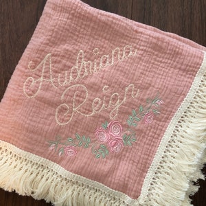 May include: A pink muslin baby blanket with white fringe trim. The blanket is embroidered with the names "Adriana Reign" and a floral design.