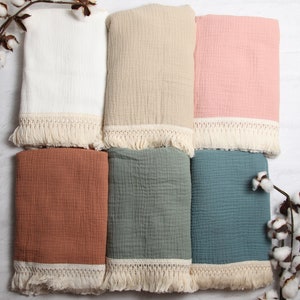 May include: Six different colored muslin baby blankets with white crochet trim and fringe. The blankets are arranged in two rows of three. The colors are white, beige, pink, rust, sage green, and teal.