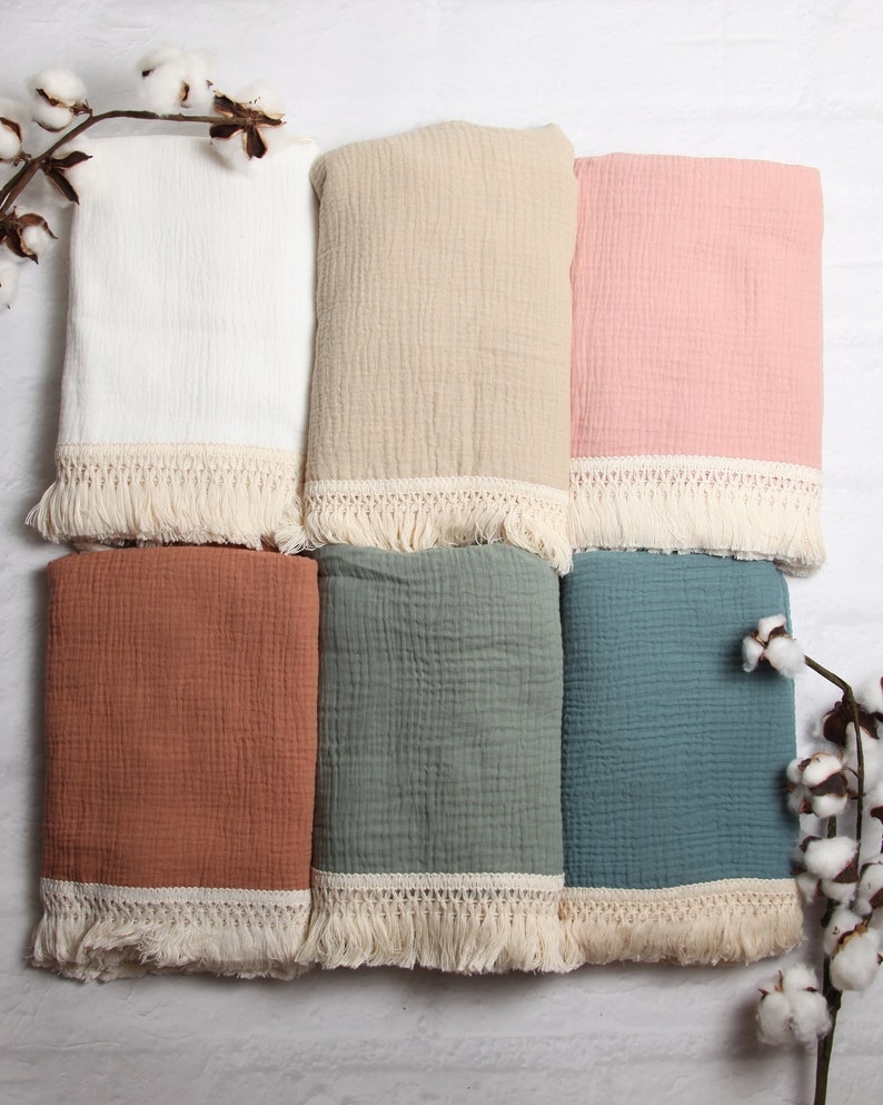 May include: Six different colored muslin baby blankets with white crochet trim and fringe. The blankets are arranged in two rows of three. The colors are white, beige, pink, rust, sage green, and teal.