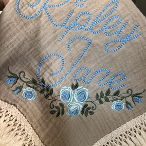 May include: A beige muslin baby blanket with a blue embroidered name "Riley Jane" and a border of blue roses and green leaves. The blanket has a white crocheted fringe around the edges.