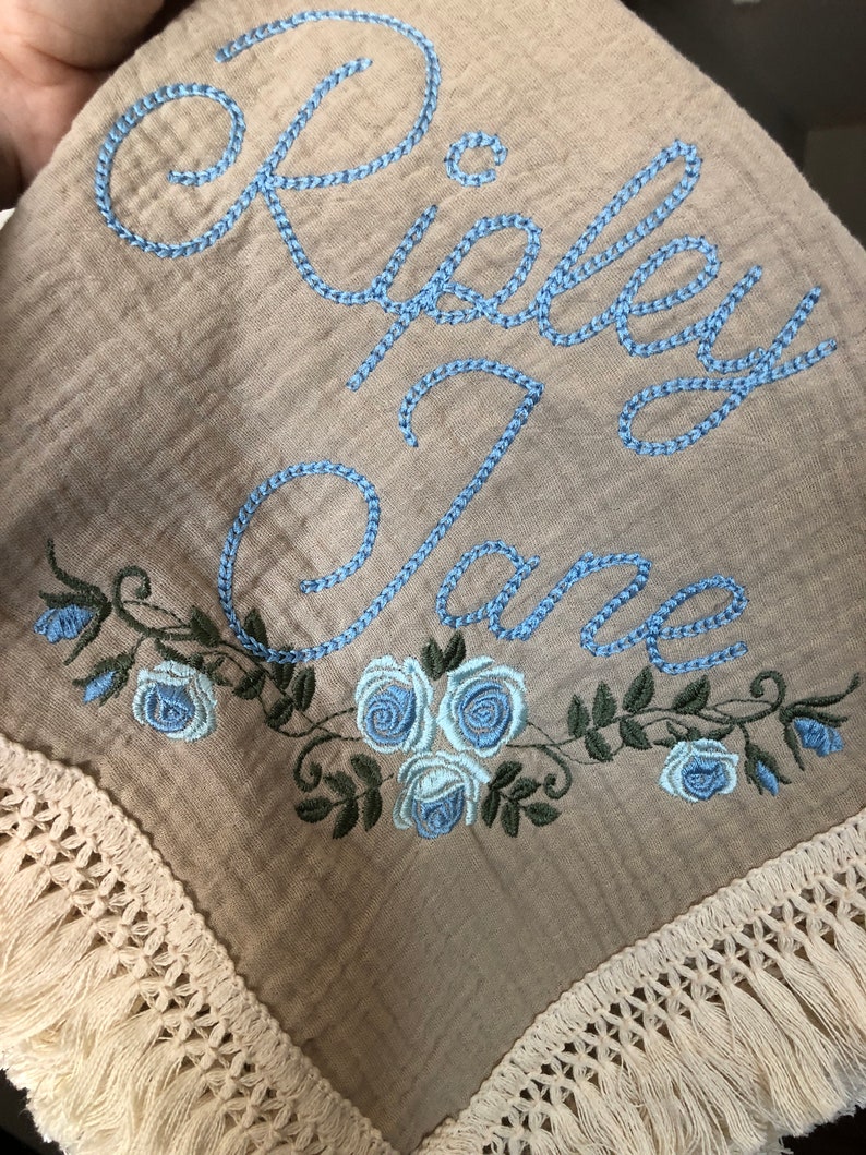 May include: A beige muslin baby blanket with a blue embroidered name "Riley Jane" and a border of blue roses and green leaves. The blanket has a white crocheted fringe around the edges.
