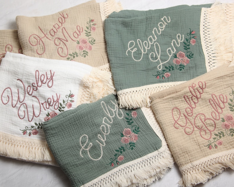 May include: Five personalized baby blankets with embroidered names and floral designs. The blankets are made of soft, white, beige, and green fabric with a fringe trim.
