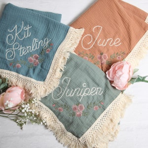 May include: Three personalized baby blankets with embroidered names and floral designs. The blankets are made of soft, lightweight fabric and have a fringe trim.