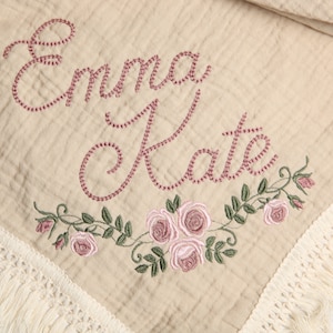 May include: A beige muslin baby blanket with a white fringe border. The blanket is embroidered with the names "Emma Kate" in pink thread and a floral design with pink roses and green leaves.