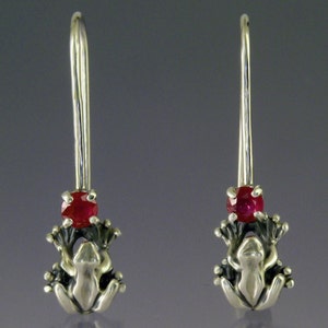 Puede incluir: Un par de pendientes de plata con forma de rana y una gema roja. Los pendientes presentan un diseño detallado de rana con un gancho largo y delgado. La gema roja está engastada sobre la cabeza de la rana. El fondo es gris.