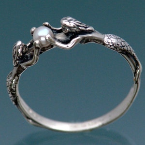 May include: A silver ring featuring two mermaids holding a pearl. The mermaids have detailed scales and flowing hair. The ring is a size 7.