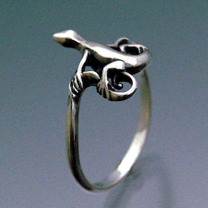 May include: A silver ring with a detailed lizard design. The lizard is perched on a branch with its tail curled around the ring band.