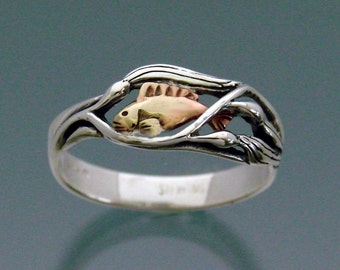 May include: A silver ring featuring a gold fish design. The fish is set within a delicate, swirling pattern reminiscent of reeds or water plants.