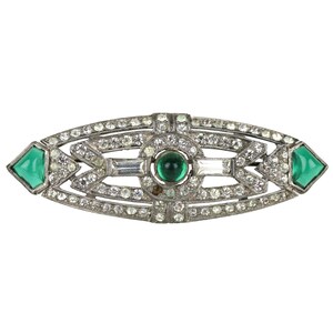 May include: A silver-toned Art Deco brooch with a central green gemstone and surrounding clear rhinestones. The brooch has a symmetrical design with green diamond-shaped accents on each end. The central design features a round green stone and baguette-cut rhinestones.