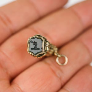 May include: A gold-toned seal with a black cameo featuring a stylised figure. The seal is attached to a gold chain with a circular ring.