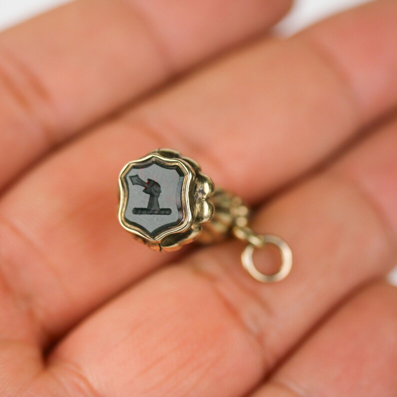 May include: A gold-toned seal with a black cameo featuring a stylised figure. The seal is attached to a gold chain with a circular ring.