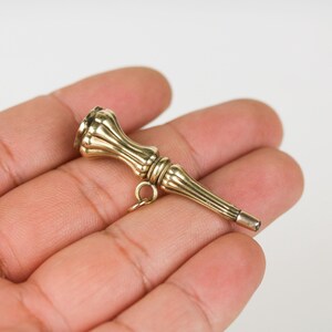 May include: A gold-toned pocket watch chain fob with a decorative, ornate design. The fob has a loop at the top for attaching to a chain and a pointed tip at the bottom.