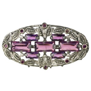 May include: Oval-shaped silver-tone brooch with a central design of rectangular and round purple gemstones. The brooch features a detailed, ornate border with leaf and berry accents, creating a vintage aesthetic. The gemstones are a vibrant shade of purple.