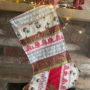 May include: A festive Christmas stocking with a patchwork design. It features patterns of holly, houses, and gold fabric. The text "celebrations" is visible. The stocking is hanging on a wooden mantelpiece above a brick fireplace.