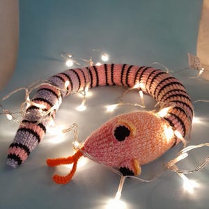May include: A crocheted snake toy in pink, black, and lavender stripes, with an orange tongue and black eyes. The snake is illuminated by a string of white fairy lights, creating a whimsical effect. The toy is on a light blue surface.