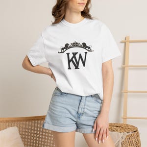 Könnte beinhalten: Weißes T-Shirt mit schwarzem Grafikdesign. Das Design zeigt eine Krone, zwei Löwen und die Buchstaben "KW". Das Shirt hat einen Rundhalsausschnitt und kurze Ärmel. Das Model trägt Jeansshorts.