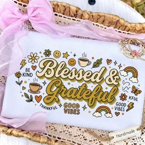 Könnte beinhalten: Ein weißes Sweatshirt mit dem Text Blessed & Grateful in einer Retro-Schriftart, umgeben von bunten Symbolen wie Kaffeetassen, Regenbögen, Smileys, Blumen, Schmetterlingen und Blitzen sowie den Schriftzügen Be Kind und Good Vibes.