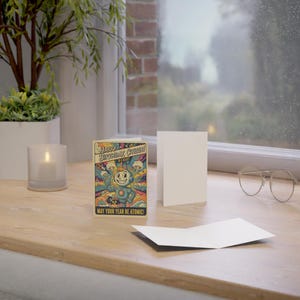 May include: A birthday card featuring a vintage cartoon robot design. The card's text reads "Happy Birthday Citizen! May Your Year Be Atomic!" It is displayed with a blank card and a lit candle on a wooden surface, near a window.