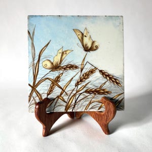 May include: A square ceramic tile featuring a painted scene of two light brown butterflies resting on stalks of golden wheat against a pale blue sky. The tile is displayed on a dark brown wooden stand with a curved, two-pronged design.