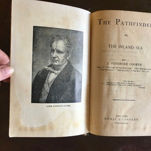 May include: A vintage book titled "The Pathfinder, or The Inland Sea" by J. Fenimore Cooper. The cover features a black and white portrait of the author, James Fenimore Cooper.