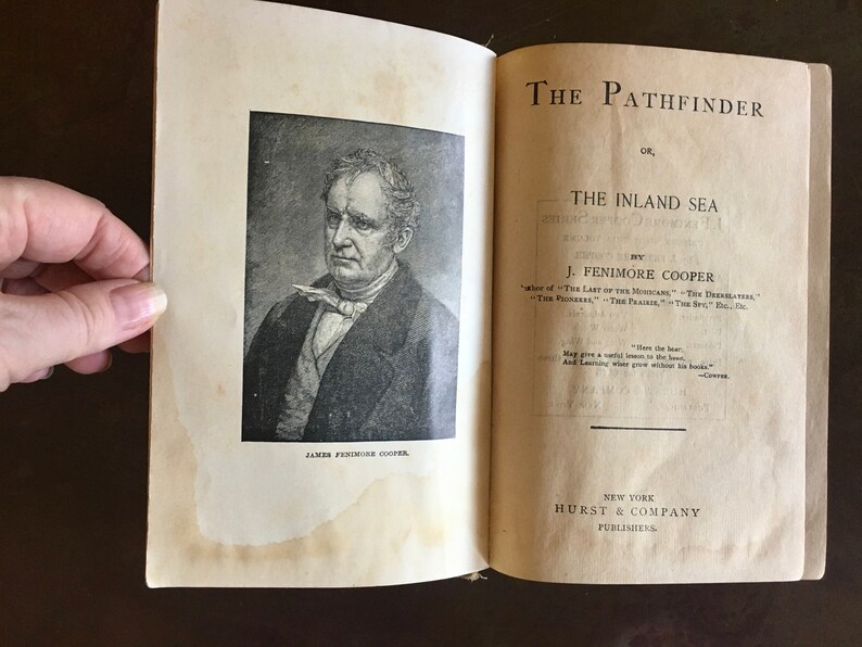 May include: A vintage book titled "The Pathfinder, or The Inland Sea" by J. Fenimore Cooper. The cover features a black and white portrait of the author, James Fenimore Cooper.