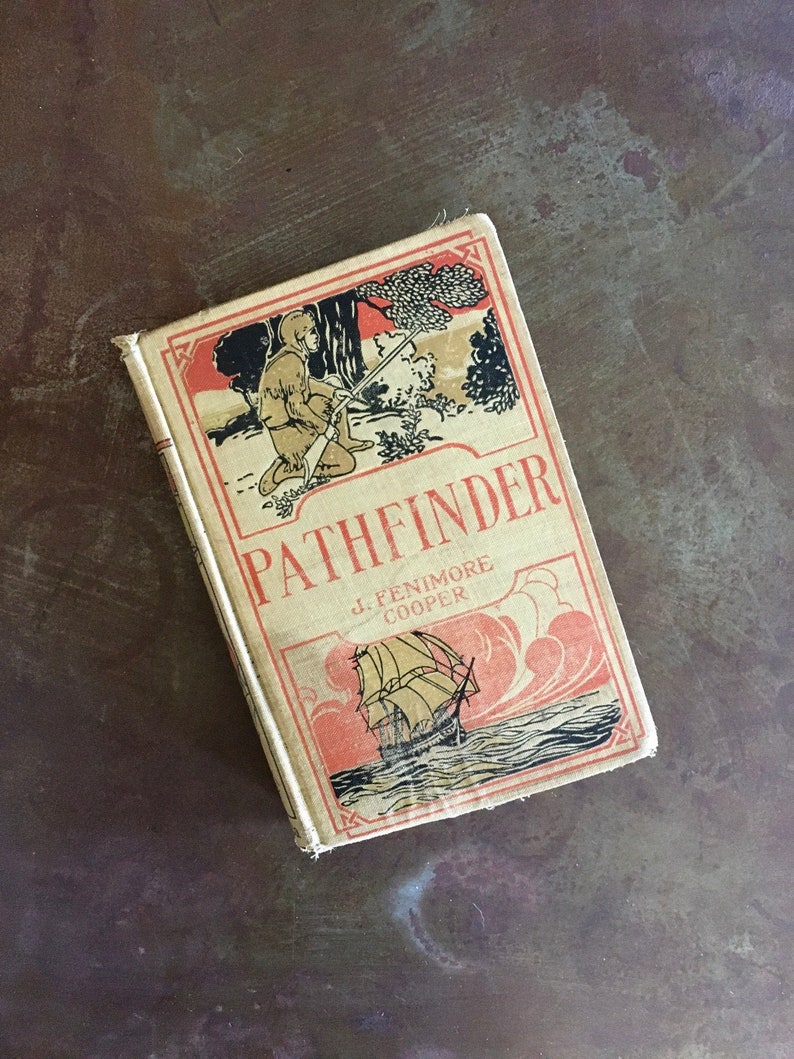 May include: A vintage book titled "PATHFINDER" by J. FENIMORE COOPER. The cover features a red and black illustration of a man in a fur hat and coat holding a rifle, standing in a forest. The bottom of the cover shows a ship sailing on the ocean.