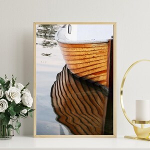 Könnte beinhalten: Ein gerahmter Druck mit einem Holzboot, das sich in ruhigem Wasser spiegelt. Der Bootsrumpf hat einen warmen Orangeton mit sichtbaren Holzplanken. Die Reflexion erzeugt ein Spiegelbild und verstärkt die visuelle Wirkung. Das Kunstwerk ist in einem hellen Holzrahmen ausgestellt.