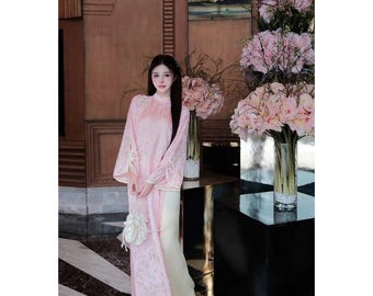 Pode incluir: Um vestido tradicional rosa claro com um padrão floral, mangas compridas e gola alta. O vestido tem detalhes brancos e é combinado com calças creme. Uma pequena bolsa branca pende do pulso. O fundo apresenta arranjos florais e uma superfície escura.