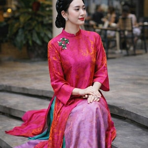 May include: A vibrant pink and orange silk dress with a high collar and long sleeves. The dress features a floral brooch and is paired with a multi-colored skirt. The woman is sitting on concrete steps.
