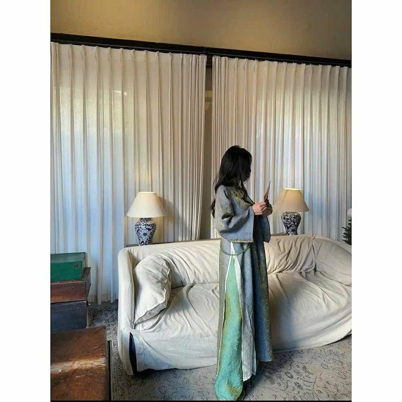 Puede incluir: Una persona con un vestido largo y fluido gris y verde con una chaqueta a juego. El vestido tiene una abertura alta y un panel verde en contraste. El fondo incluye un sof&aacute; blanco, cortinas y l&aacute;mparas.