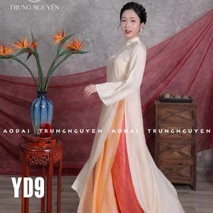 Könnte beinhalten: Eine Frau trägt ein cremefarbenes Ao Dai-Kleid mit hohem Kragen und langen Ärmeln. Das Kleid hat einen geteilten Rock, der eine pfirsich- und orangefarbene Unterschicht freilegt. Das Bild enthält eine Vase mit orangefarbenen Blumen und den Text "TRUNG NGUYEN" und "AODAI".