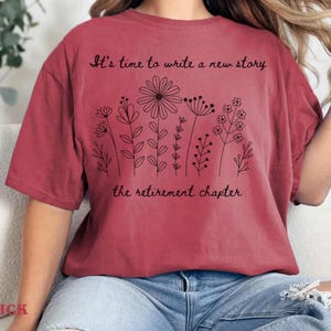 May include: A brick red short-sleeved t-shirt featuring a black line art illustration of various wildflowers. The text "It's time to write a new story" is printed above the flowers, and "the retirement chapter" is printed below the floral design.