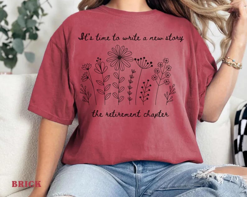 May include: A brick red short-sleeved t-shirt featuring a black line art illustration of various wildflowers. The text "It's time to write a new story" is printed above the flowers, and "the retirement chapter" is printed below the floral design.