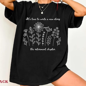May include: A black short-sleeved t-shirt featuring a white line-art graphic of various wildflowers. The text "It's time to write a new story" is printed above the flowers, and "the retirement chapter" is printed below the floral design.