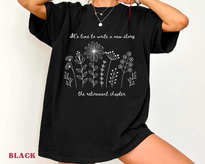 May include: A black short-sleeved t-shirt featuring a white line-art graphic of various wildflowers. The text "It's time to write a new story" is printed above the flowers, and "the retirement chapter" is printed below the floral design.