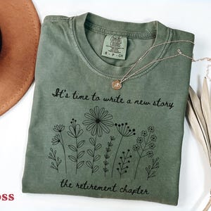 May include: A moss green short-sleeve t-shirt featuring a black line-art graphic of various wildflowers. The text reads, "It's time to write a new story, the retirement chapter." The shirt has a crew neckline and a Comfort Colors brand label.