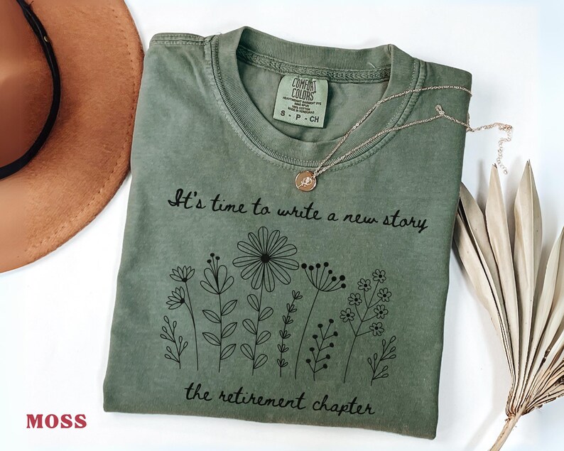 May include: A moss green short-sleeve t-shirt featuring a black line-art graphic of various wildflowers. The text reads, "It's time to write a new story, the retirement chapter." The shirt has a crew neckline and a Comfort Colors brand label.