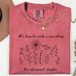 May include: A watermelon-colored t-shirt featuring a black line-art illustration of various wildflowers. The text reads, "It's time to write a new story" above the flowers and "the retirement chapter" below. The shirt has a Comfort Colors label.