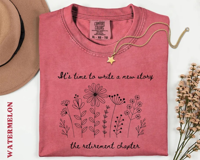 May include: A watermelon-colored t-shirt featuring a black line-art illustration of various wildflowers. The text reads, "It's time to write a new story" above the flowers and "the retirement chapter" below. The shirt has a Comfort Colors label.