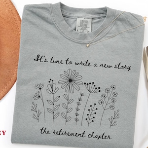 May include: A grey short-sleeved t-shirt featuring a black line art illustration of various wildflowers. The text "It's time to write a new story" and "the retirement chapter" is printed in black script above and below the floral design.