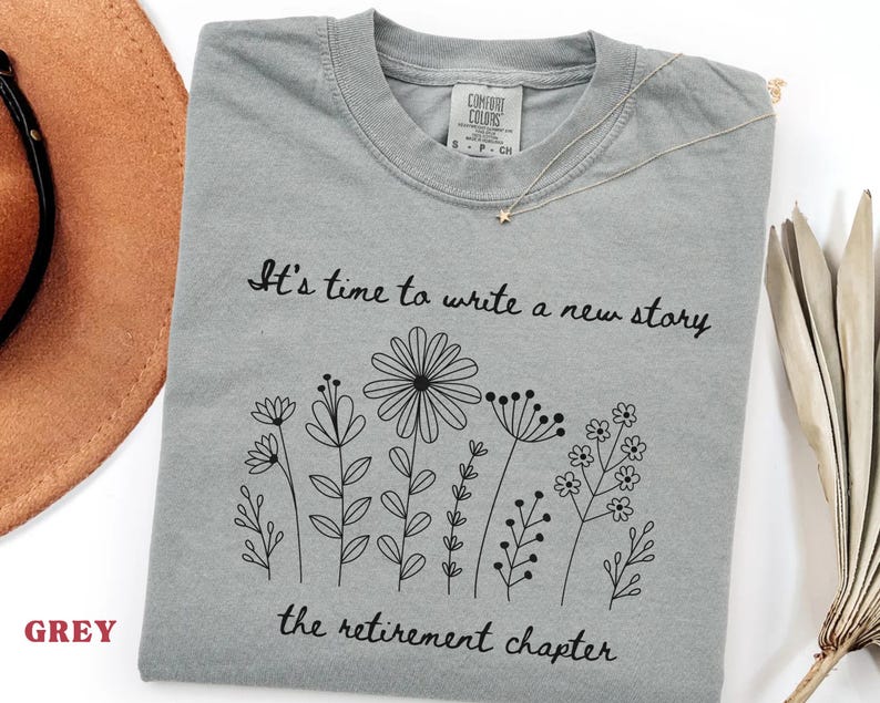 May include: A grey short-sleeved t-shirt featuring a black line art illustration of various wildflowers. The text "It's time to write a new story" and "the retirement chapter" is printed in black script above and below the floral design.