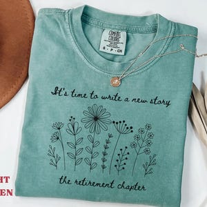 May include: A light green short-sleeved t-shirt featuring a black line art graphic of various wildflowers. The text reads, "It's time to write a new story" above the flowers and "the retirement chapter" below the flowers in a cursive font.